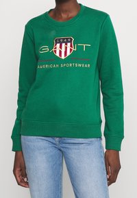 Groene sweatshirt van een katoenmix met ribbels aan de mouwen en de zoom. Voorzien van een geborduurd logo in geel, rood en marineblauw. Klassieke ronde halslijn.