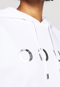 Sudadera blanca con letras plateadas reflectantes. Cuerdas con puntas metálicas en el cuello. Textura de tela suave y lisa.