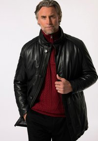 Černá kožená bunda s vysokým límcem, zapínáním na zip a předními knoflíky, nošená přes červený strukturovaný svetr. Hladký povrch a dobře padnoucí střih.