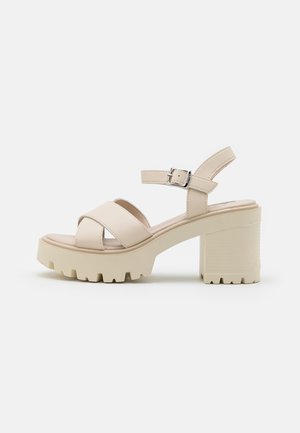 Beige sandal med chunky hæl og tyk sål, krydsede frontremme og justerbar ankelrem med spænde.