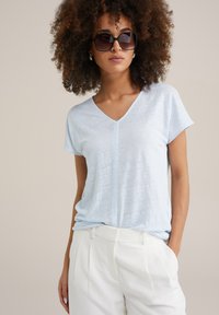 Jasnoniebieska koszulka typu V-neck z lekkiego, teksturowanego materiału, zestawiona z białymi dopasowanymi spodniami. Brak widocznych wzorów lub ozdobników.