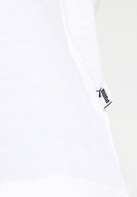 Biała bawełniana koszulka z krótkim rękawem, z czarną metką z logo Puma na dole. Gładka faktura, prosty i czysty design.