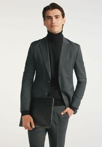 Szary żakiet i spodnie z teksturowanej tkaniny, w połączeniu z czarnym golfem. Trzyma elegancki czarny skórzany pokrowiec na laptopa.