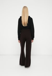 Femme blonde de dos, portant un sweat à capuche noir, un pantalon large marron foncé et des baskets blanches, sur fond blanc uni.