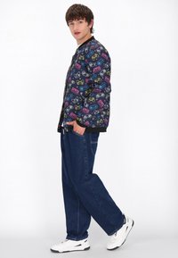 Giacca bomber nera con grafiche colorate a tema videogioco, abbinata a jeans in denim blu dal taglio largo e sneakers bianche.