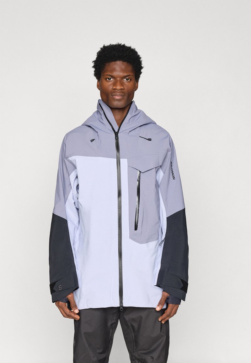 Άνδρας που φοράει ένα ανοιχτό μωβ και μαύρο μπουφάν με κουκούλα, με φερμουάρ και θ pockets της βέλκρο, στέκεται απέναντι σε ένα απλό ανοιχτό φόντο.