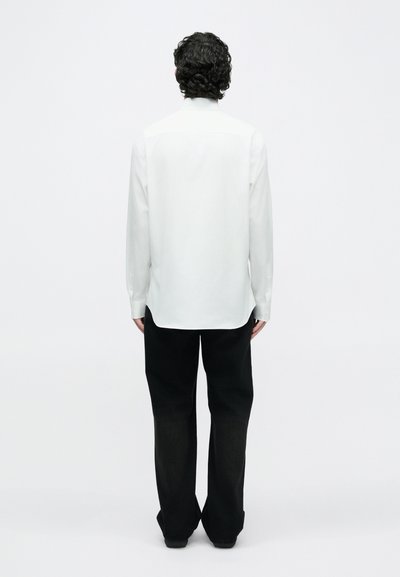 Mand med krøllet mørkt hår står med ryggen til iført en hvid langærmet skjorte og løse sorte bukser mod en ensfarvet hvid baggrund.