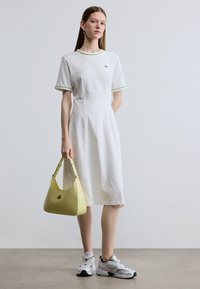 Λευκό βαμβακερό φόρεμα με πράσινη διακόσμηση, κοντά μανίκια και άνετη εφαρμογή. Κίτρινη τσάντα χειρός με στρογγυλεμένο σχήμα και λεπτομέρεια κοψίματος. Γκρι αθλητικά παπούτσια.