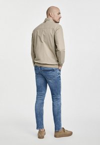 Beżowa, lekka kurtka z stójką, dopasowanym talią i ribbowym wykończeniem, zestawiona z jasnoniebieskimi, slim fit dżinsami i brązowymi butami.