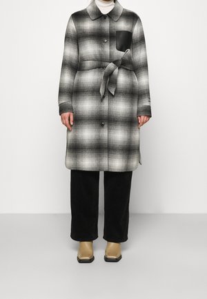 Personne portant un manteau ceinturé à carreaux gris et noir avec une poche poitrine noire, un pantalon noir, des bottes beiges à bout carré et un col roulé blanc.