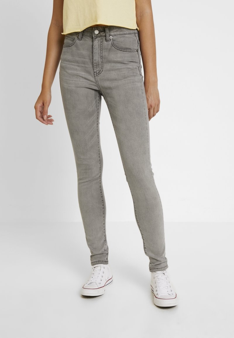 Jeans gris de cintura alta con un ajuste slim, que cuentan con bolsillos frontales, pasadores para cinturón y una textura de denim suave. Llevados con zapatillas blancas.