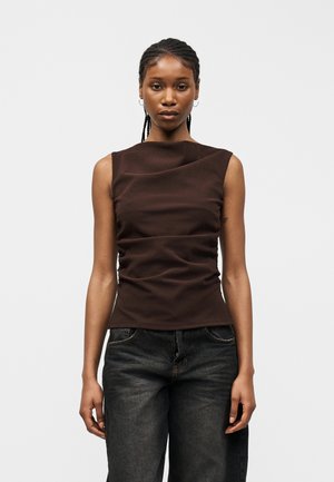 Γυναίκα με πλεξούδες, φορούσα αμάνικο σκουρό καφέ τοπ με ρυτιδώσεις και φαρδιά μαύρα τζιν, στέκεται μπροστά σε απλό ανοιχτό φόντο.