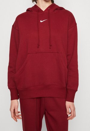 Osoba ubrana w czerwoną bluzę z kapturem Nike i pasujące czerwone dresy, z rękami swobodnie opartymi wzdłuż boków.