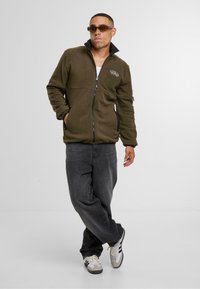 Oliivinvihreä fleece-takki korkealla kauluksella, vetoketjusuljenta ja brodeerattu yksityiskohta. Yhdistetty tummanharmaiden farkkujen ja valkoisten urheilukenksien kanssa.