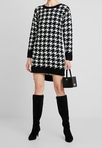 Černobíl dress s vzorem houndstooth, kulatým výstřihem, dlouhými rukávy a bočními rozparky, v kombinaci s černými kozačkami po kolena a kabelkou.