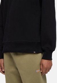 Svart sweatshirt med ribbad nederkant och muddar, tillsammans med olivgröna byxor som har en cirkulär logotypapplikation.