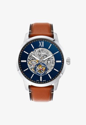 Montre ronde argentée Fossil avec cadran bleu, engrenages mécaniques exposés, index blancs et bracelet en cuir marron.