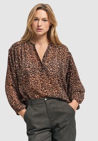Luipaardprint blouse van doorschijnende bruine stof, met een knoopsluiting bij de v-hals en lange mouwen, gecombineerd met grijze, op maat gemaakte broek.