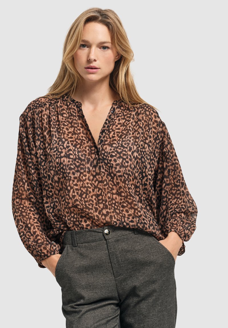 Luipaardprint blouse van doorschijnende bruine stof, met een knoopsluiting bij de v-hals en lange mouwen, gecombineerd met grijze, op maat gemaakte broek.