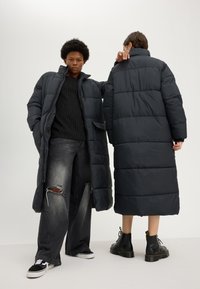 Two people wear long black puffer coats; one faces forward with hands in pockets, the other stands back wearing black boots and ripped jeans.