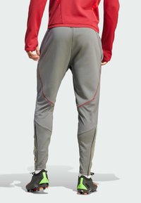 Pantalones deportivos grises con un diseño ajustado, con costuras rojas y puños con cremallera. Llevados con botas de fútbol negras con detalles en verde.