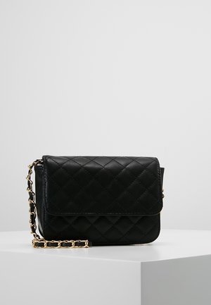 Sac bandoulière en cuir noir matelassé avec une chaîne dorée, posé sur un socle blanc devant un fond gris uni.