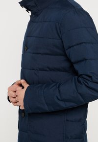 Marinblå puffjacka med vertikala sömmar, utrustad med ståkrage, knapplist och puffig struktur på kroppen och ärmarna.