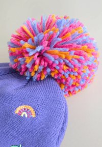 Bonnet en laine tricotée violet avec un pompon coloré comportant des fils roses, bleus et orange ; détail décoratif brodé en arc-en-ciel sur le côté.
