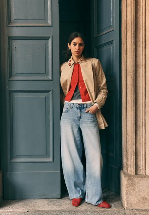 Ženska, ki stoji v vratih, oblečena v bež trenč plašč, rdeč kardigan, sivo majico, široke svetlo modre kavbojke in rdeče čevlje.