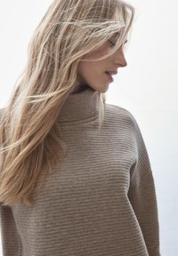 Beige ribbad turtleneck-pusero, jossa on rento malli. Pehmeä rakenne ja kevyet kankaat, joissa on korkea kaulus ja minimalistinen muotoilu.
