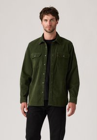 Green corduroy shirt with button closure, two chest pockets, and a classic collar, paired with black jeans. Smooth texture and structured design.