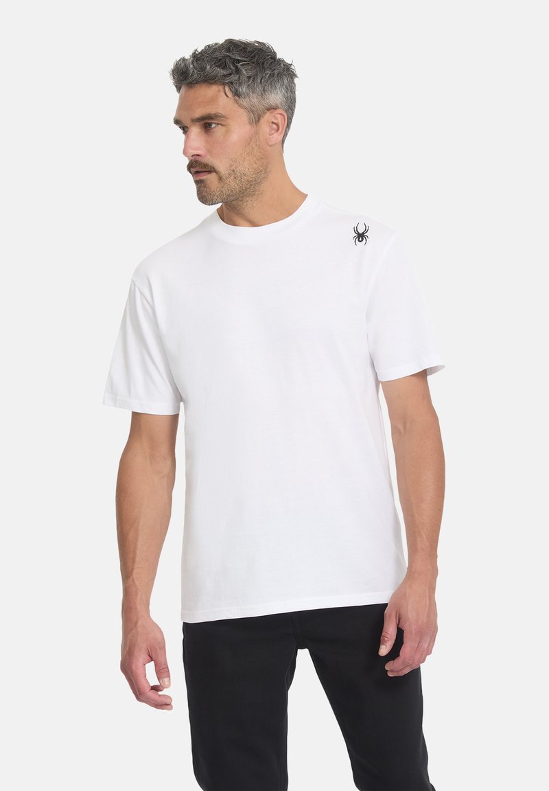 Homme aux cheveux gris portant un t-shirt blanc uni à manches courtes avec un petit logo noir d’araignée sur l’épaule gauche et un pantalon noir.