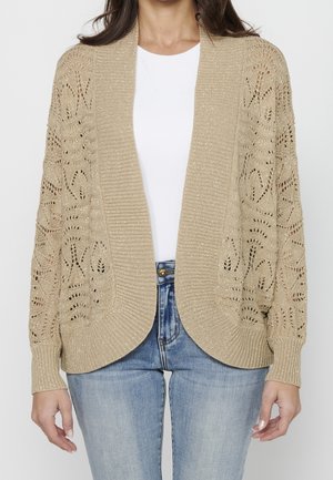Beige strikket cardigan med åben front, tekstureret mønster på ærmerne, ribbede kanter og metalltråd accenter. Båret over en hvid top.