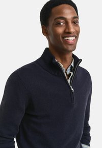 Navy Zip-Up-Pullover mit hohem Rippenkragen, aus weichem Stoff gefertigt. Besitzt einen silbernen Reißverschluss und kragenunterhalb eine geschichtete Hemdkragen.
