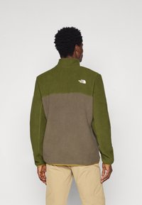 Giacca in pile con colletto alto, caratterizzata da pannelli verde scuro e verde oliva, texture morbida e un logo bianco sulla parte superiore della schiena.