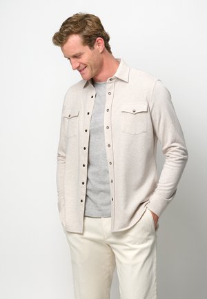 Hellbeige Hemdbluse mit Kragen, zwei Brusttaschen und langen Ärmeln, getragen über einem grauen T-Shirt, kombiniert mit cremefarbenen Hosen.