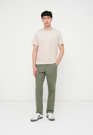 Άνδρας που στέκεται μπροστά σε λευκό φόντο, φορώντας μπεζ μπλούζα, παντελόνι ελιάς και λευκά αθλητικά με μαύρες ρίγες, με τα χέρια χαλαρά.