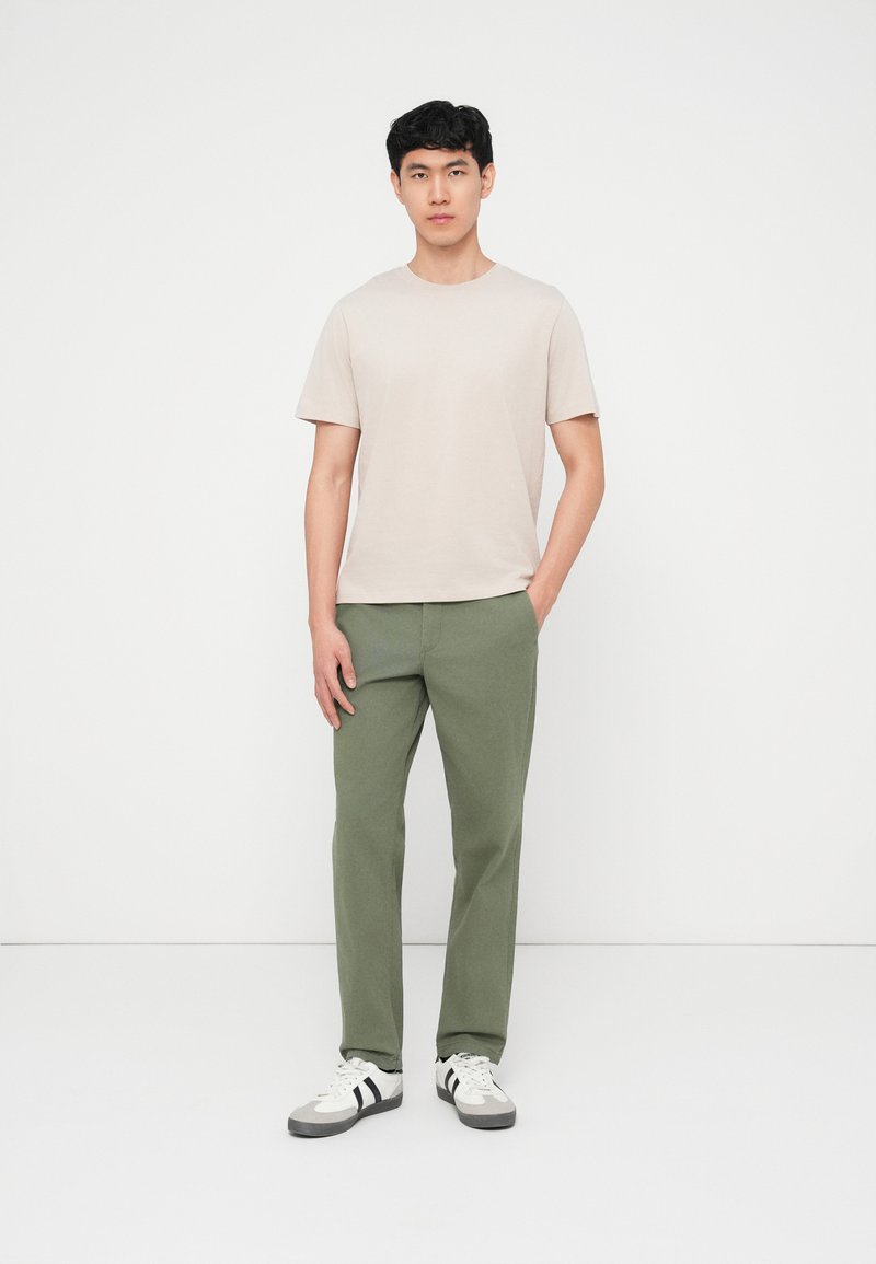 Mann stående mot hvit bakgrunn iført beige t-skjorte, olivengrønne bukser og hvite joggesko med svarte striper, hendene avslappet.