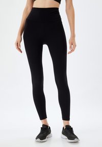 Hooggewaisted zwarte legging gemaakt van rekbaar materiaal, met een gladde textuur en een taps toelopend ontwerp, gecombineerd met donkere sportschoenen.