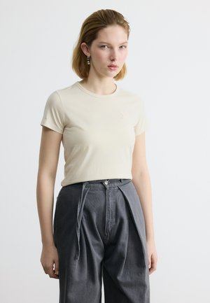 Νεαρή γυναίκα που φοράει κρεμ κοντομάνικη μπλούζα και ψηλόμεσο σκούρο πλισέ παντελόνι στέκεται μπροστά σε ένα απλό φωτεινό φόντο.