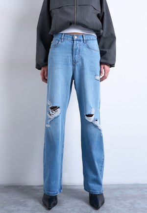 Personne portant un jean déchiré bleu clair, des bottes pointues noires et une veste gris foncé à fines rayures, debout contre un mur uni.