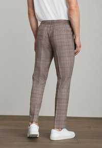 Brune ternede bukser med elastisk talje, slim fit og sidelommer, parret med hvide sneakers på et trægulv.