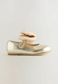 Chaussure Mary Jane métallique dorée avec bride de cheville beige douce à volants et semelle plate sur fond clair.