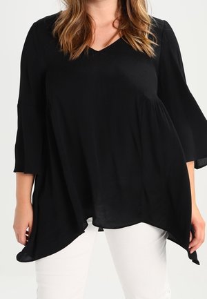 Vrouw draagt een losse zwarte tuniek met V-hals, driekwartmouwen en asymmetrische zoom, gecombineerd met een witte broek, staand tegen een effen achtergrond.