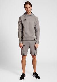 Grå hoodie och matchande shorts i mjukt tyg. Har svart logotyp och sidopaneler. Modellen står i träningsskor mot en neutral bakgrund.