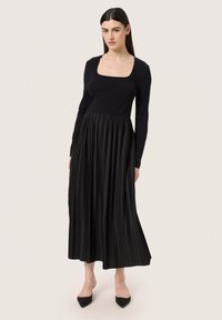 Black dress with a fitted long-sleeve top and a pleated skirt. Features a square neckline, smooth texture, and ankle-length design.