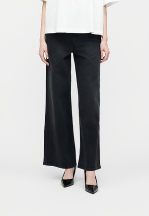 Person trägt schwarze Jeans mit weitem Bein und schwarze spitze Absätze, kombiniert mit einem weißen kurzärmeligen Oberteil vor einem einfarbigen Hintergrund.