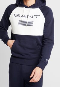 Sudadera azul marino con panel frontal blanco que presenta el logo "GANT" y rayas. Tejido suave, cordón ajustable y bolsillo tipo canguro.