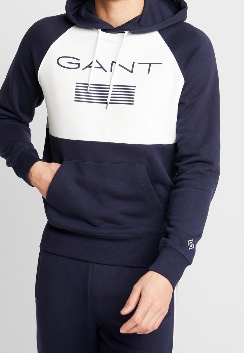 Sudadera azul marino con panel frontal blanco que presenta el logo "GANT" y rayas. Tejido suave, cordón ajustable y bolsillo tipo canguro.