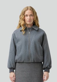 Una donna con lunghi capelli mossi indossa una giacca bomber grigia su una gonna a scacchi, posando su uno sfondo grigio uniforme.